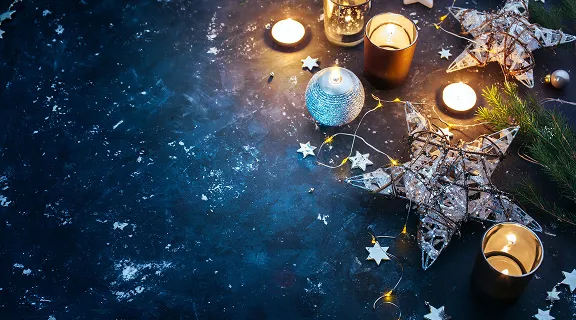 Dunkler, blauer Hintergrund mit Weihnachtsdekoration, funkelnden Lichterketten, sternförmigen Lichtern und brennenden Teelichtern.